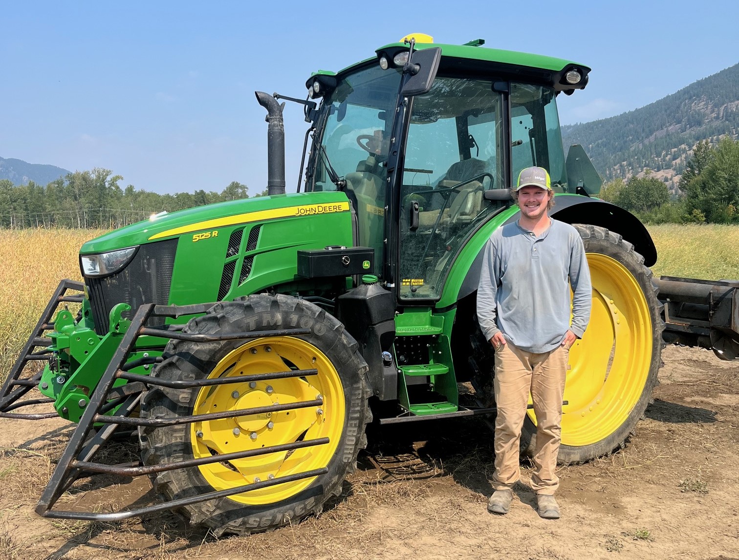 A smiling man in a grey shirt and cap stands in a field beside a green John Deere tractor.