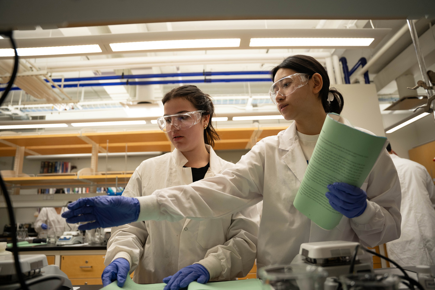 Two students in white lab coats, safety goggles, and blue gloves work together at a laboratory bench.