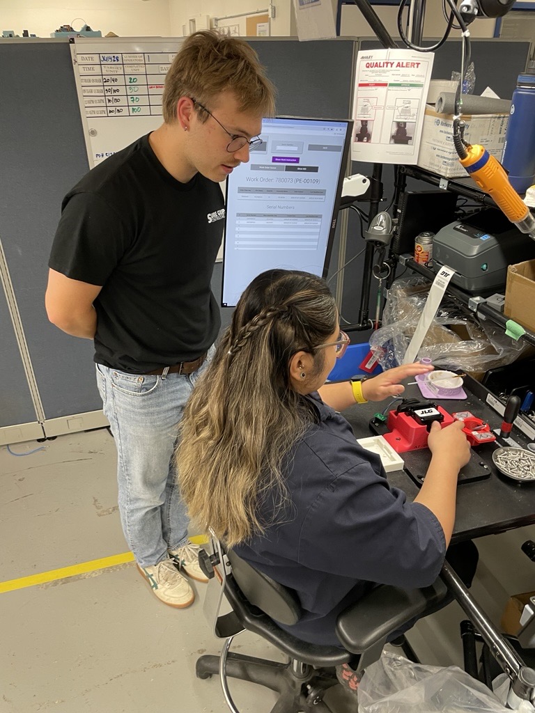 A supervisor watches a co-op student work at an assembly station.
