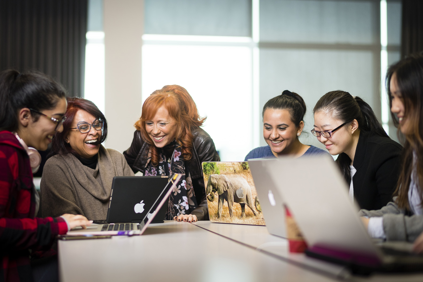 A group of smiling students and a mentor working together on laptops at a table.