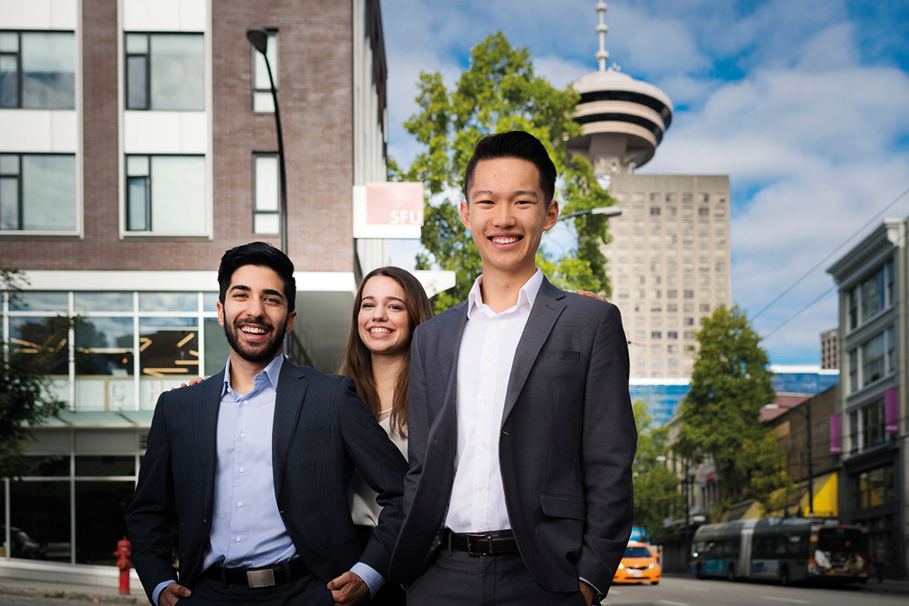 Three professionally dressed students stand together on a city street, with a distinctive tower and urban buildings visible in the background.