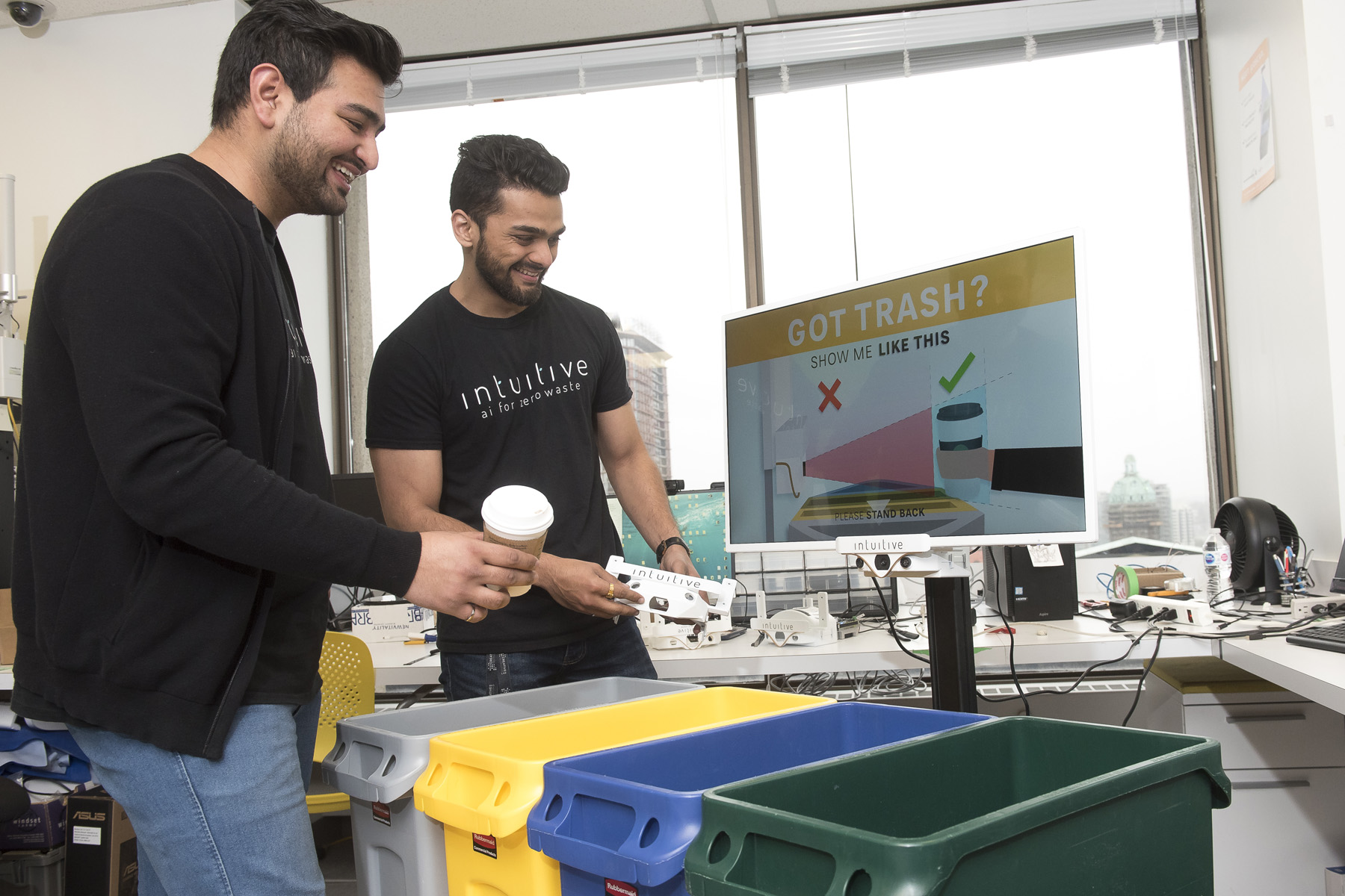 Two people sort waste near colour-coded recycling bins in a laboratory or workshop. A sign behind them promotes proper waste sorting.