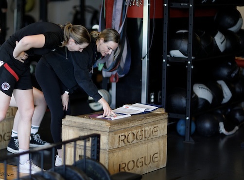 Two people organize materials at a table inside a sports facility, surrounded by shelves of sports equipment and protective gear.