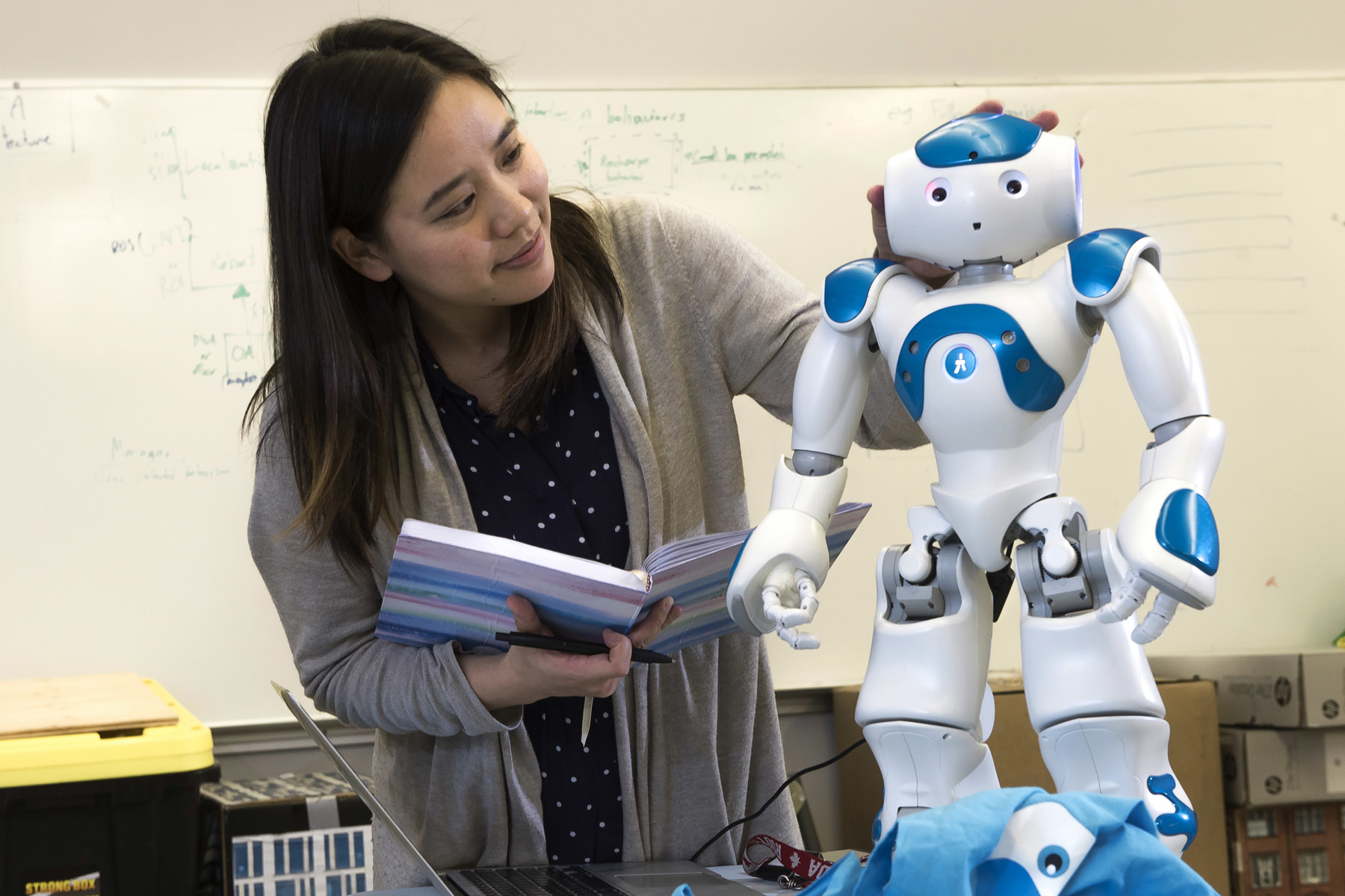 A woman holds a notebook beside a small humanoid robot in a classroom or laboratory, with a whiteboard in the background.
