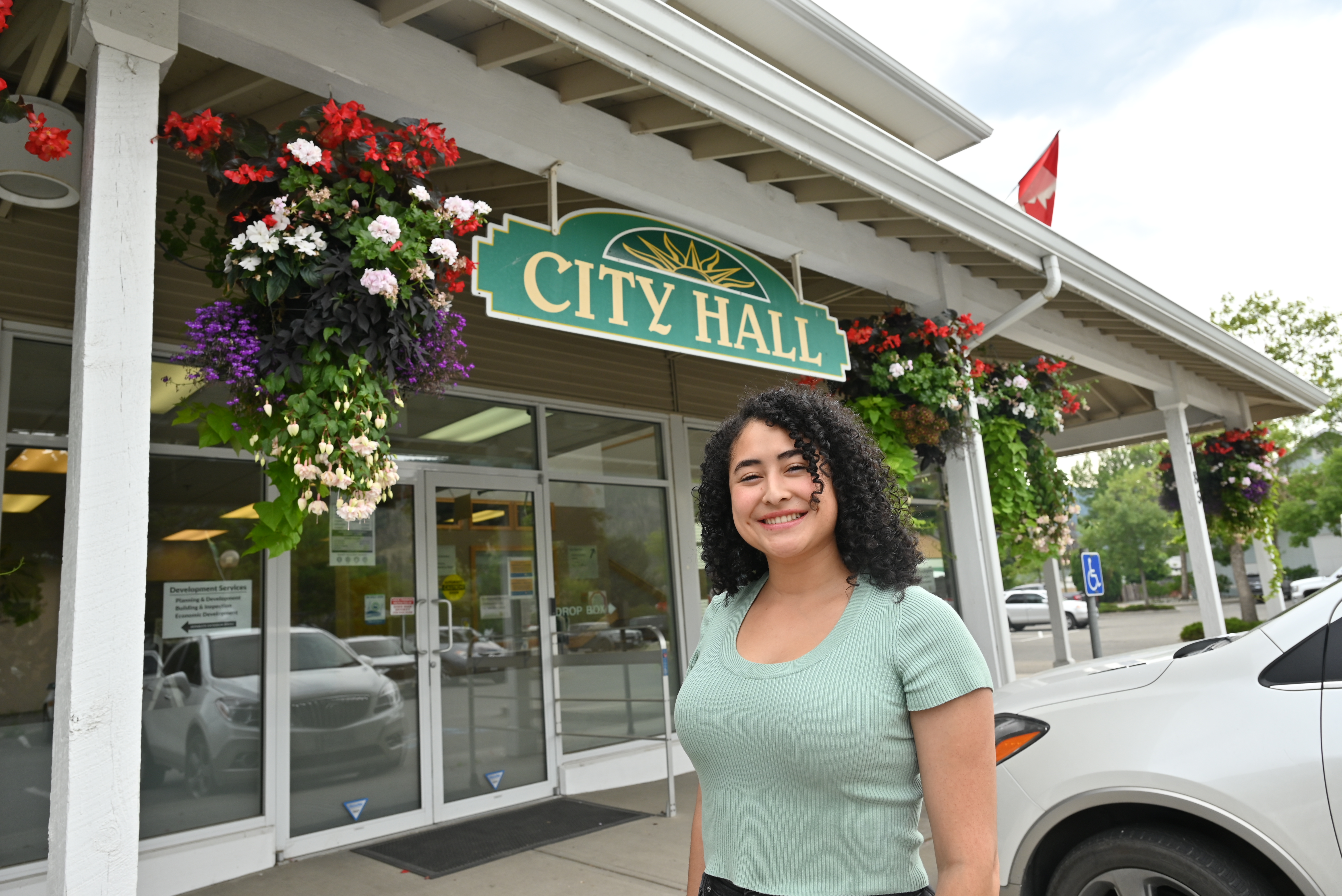 A smiling woman stands outside a city hall building decorated with colorful hanging flower baskets.