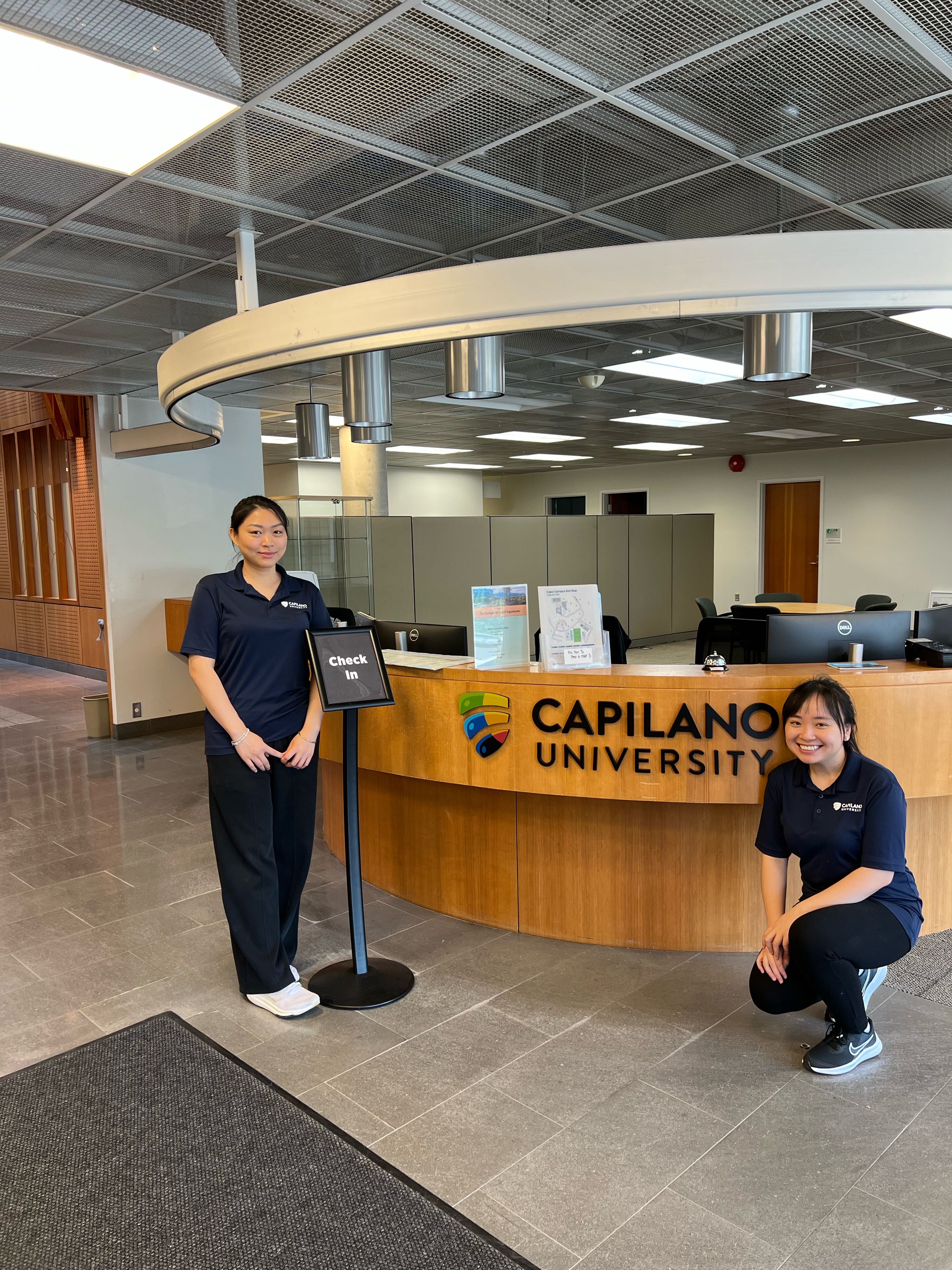 Two university staff members pose at the reception desk inside Capilano University.