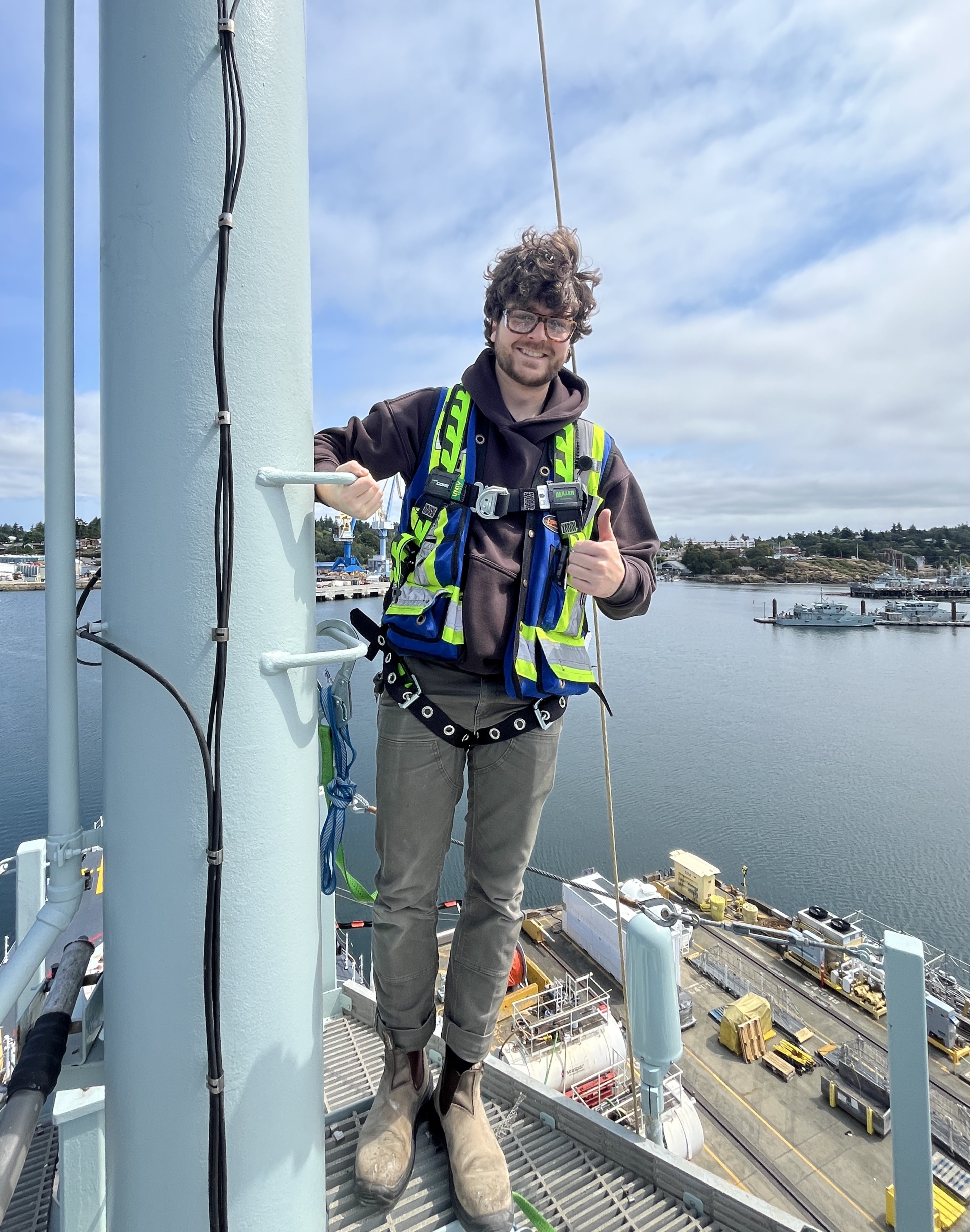 A worker in safety gear stands on an industrial tower overlooking a harbor.