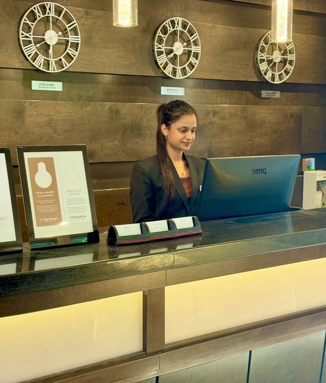 A hotel receptionist stands behind a modern front desk with a computer, framed awards, and three large wall clocks.